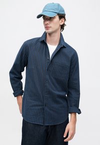 Joven vestido con una camisa de botones a rayas azules, camiseta blanca debajo, jeans azules y una gorra de béisbol de mezclilla azul claro, mirando hacia un lado.