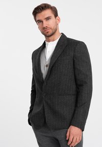 Blazer a righe grigie realizzato in tessuto strutturato, dotato di chiusura a un bottone, rever a punta e taschino sul petto. Indossato sopra una camicia bianca.