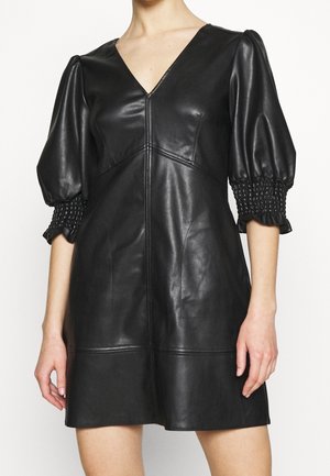 Kobieta w krótkiej czarnej sukience z ekoskóry z dekoltem w serek oraz bufkami i marszczonymi rękawami o długości do połowy przedramienia.