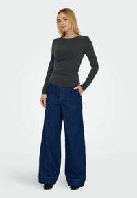 Langærmet mørkegrå bluse med sammensyning, parret med brede mørkeblå denimjeans med synlige stikninger og forlommer.