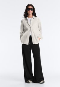 Femme portant un pantalon noir à jambes larges, des baskets blanches, un haut blanc, une chemise beige à carreaux boutonnée et des lunettes de soleil noires, debout les mains dans les poches.