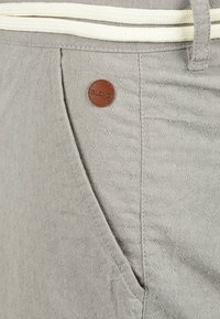 Shorts gris clair en tissu texturé, dotés d'une ceinture crème contrastante et d'un patch circulaire marron avec l'inscription "BLEND."