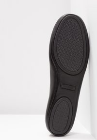Semelle de chaussure habillée en cuir noir avec motifs en caoutchouc texturés antidérapants sur les zones du talon et des orteils, présentée verticalement sur fond blanc.
