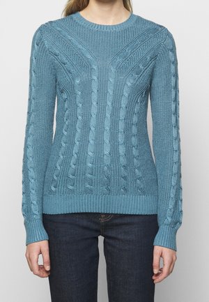 Kvinde iført en tætsiddende lyseblå kabelstrikket sweater med lange ærmer og mørkeblå jeans, stående mod en ensartet lys baggrund.