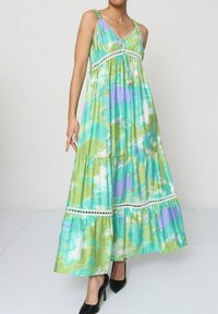 Robe maxi en tie-dye vert et bleu avec des bordures en dentelle, un ourlet en volants et des bretelles spaghetti. Tissu lisse, silhouette fluide, portée avec des talons noirs.