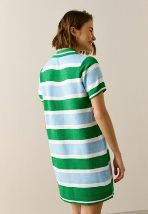 Naeratav naine, kandes lühikeste varrukatega kootud kleiti, millel on laiad horisontaalsed rohelised, sinised ja valged triibud, seistes ühevärvilise heleda tausta ees.