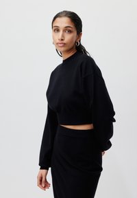 Svart cropped genser med lange ermer og rund hals, laget av glatt stoff. Brukt med matchende høyhalsede skjørt.