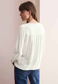 Blouse blanche à manches longues avec une texture douce, présentant un détail plissé au dos et un col rond, associée à un jean en denim bleu.
