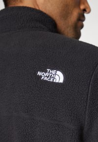 Svart fleecejakke med høy krage og en hvit brodert logo av "The North Face" på venstre øvre rygg. Myk tekstur.