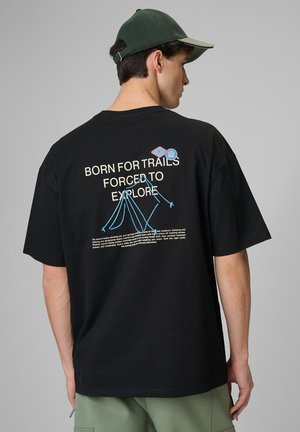 Mężczyzna w czarnej koszulce z niebieską górską linią i napisem „Born for trails forced to explore” oraz zieloną czapką, odwrócony tyłem.