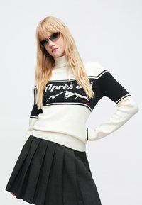 Blond kvinne med mørke solbriller, hvit og svart genser med fjellmotiv og teksten "Après Ski", og en svart plissert skjørt.