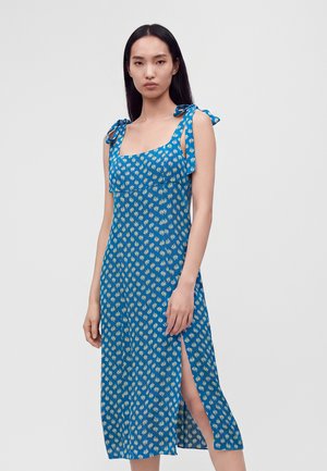 Femme aux longs cheveux noirs portant une robe bleue sans manches avec des motifs de feuilles blanches et une fente latérale, debout devant un fond clair uni.