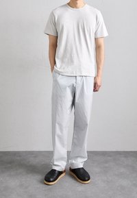 T-shirt grigio chiaro a maniche corte, pantaloni a righe blu chiaro e zoccoli neri con suola di legno, in piedi contro uno sfondo neutro.