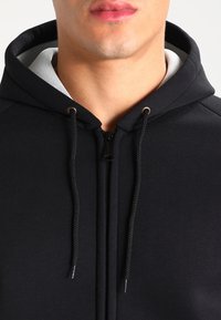 Sudadera negra con cremallera en el frente, cordones y un forro interior de capucha en un tono gris claro que contrasta. Tela suave y texturizada.