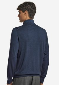 Man som bär en marinblå långärmad tröja med hög krage, sedd bakifrån mot en vit bakgrund.