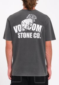 Camiseta de algodón gris con un gran gráfico blanco de una calavera y el texto "VOLCOM STONE CO." en la parte trasera. Mangas cortas, cuello redondo.