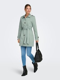 Trench-coat vert clair avec fermetures à boutons et ceinture à nouer, porté avec un jean skinny bleu et des bottines noires. Porte un sac noir.