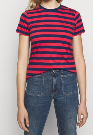 Persoon draagt een rood- en marineblauw gestreept shirt met korte mouwen, gecombineerd met hooggetailleerde blauwe jeans met voorzakken.