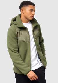 Fleece-zip-Jacke in olivgrün mit einem gepolsterten Brustpaneel. Verfügt über eine Kapuze, Fronttaschen und kontrastierende Stoffstrukturen.