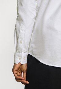 Levá paže osoby, která má na sobě bílou košili s dlouhým rukávem s knoflíkovými manžetami a černé kalhoty, na jednobarevném pozadí.