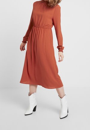 Woman wearing a rust-colored long-sleeve midi dress paired with white mid-calf heeled boots, standing against a plain background.