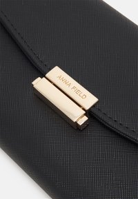 Black textured clutch featuring a gold metallic clasp with embossed "ANNA FIELD" logo, emphasizing sleek design and sophisticated materials.