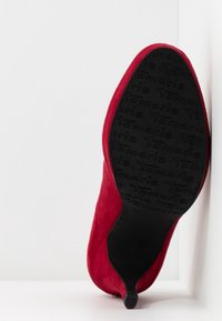 Chaussure en daim rouge avec un bout pointu et une fine semelle en caoutchouc noire présentant une surface texturée avec le nom de la marque "Tamaris" embossé.