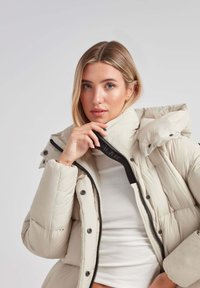 Beige puffer jakke med høj krave, lynlåslukning og trykknapper. Har en quiltet tekstur og sorte metaldele.