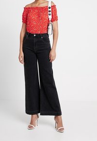 Kvinne iført en rød blomstret topp med avdekkede skuldre, høy midje brede svarte jeans, hvite høyhælte sandaler og en hvit skulderveske.