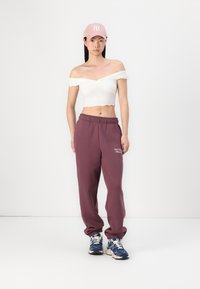 Bijela crop bluza s otkrivenim ramenima i valovitim rubovima, u kombinaciji s burgundskim trenirkama koje imaju izvezeni logo i plave tenisice.