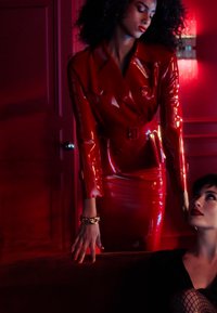 Manteau en latex rouge, brillant, à double boutonnage, porté sur une jupe crayon assortie, rehaussé par un bracelet en chaîne dorée ; fond sombre avec un éclairage rouge.