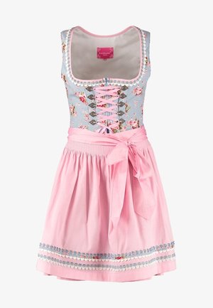 Młoda kobieta ubrana w tradycyjną kwiecistą suknię dirndl z białą bluzką, różowym fartuszkiem wiązanym w talii, białymi rajstopami i beżowymi sznurowanymi butami.