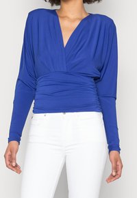 Blå långärmad topp med djup V-ringning, samlad midja och slät textur, kombinerad med vita åtsittande jeans.