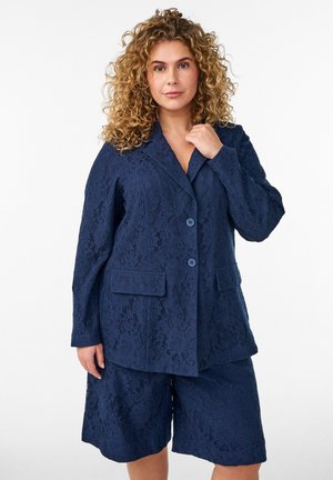 Kvinde med krøllet blondt hår iført en marineblå blondeblazer og matchende knæhøje shorts, poserer med den ene hånd nær kraven.