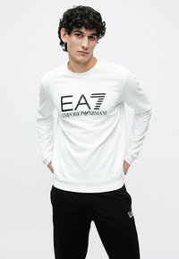 Fiatal férfi göndör hajjal, fehér EA7 Emporio Armani hosszú ujjú ingben és fekete nadrágban, kezeit a háta mögött tartva áll.