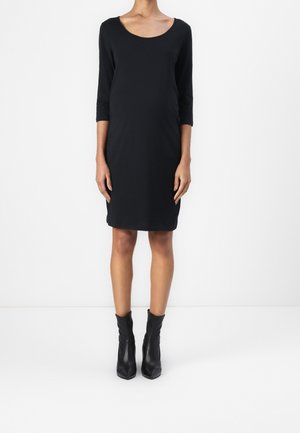 Frau in einem schwarzen knielangen Kleid mit Dreiviertelärmeln und schwarzen Stiefeletten, stehend vor einem schlichten weißen Hintergrund.