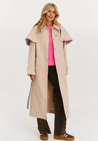 Žena v béžovém dlouhém trenčkotě, jasně růžovém svetru, kalhotách s leopardím potiskem a hnědých teniskách s růžovými pruhy, stojící na bílém pozadí.