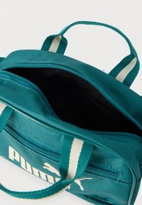 Τσαντάκι Teal Puma με λευκό λογότυπο, δύο λαβές με λευκές ρίγες και ανοιχτό φερμουάρ που αποκαλύπτει την μαύρη εσωτερική επένδυση με την επιγραφή Puma.