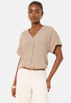 Woman wearing beige short-sleeve V-neck blouse with rolled cuffs and white high-waist pants, accessorized with a brown belt and gold hoop earrings.