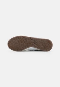 De buitenzool van de sportschoen heeft een gestructureerd bruin rubberpatroon met ronde grip, in contrast met een witte bovenkant met grijze accenten.
