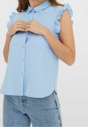 Femme boutonnant une blouse sans manches bleu clair avec des épaules volantées, associée à un jean denim bleu clair.