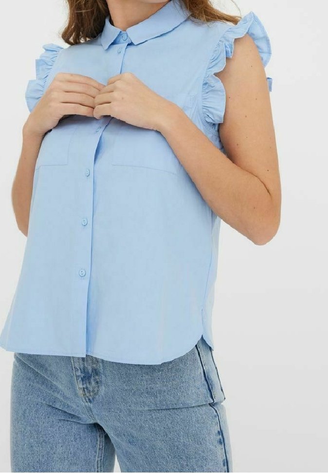 Femme boutonnant une blouse sans manches bleu clair avec des épaules volantées, associée à un jean denim bleu clair.