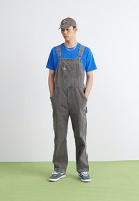 Salopette grise avec poches, portée par-dessus un t-shirt bleu. Une casquette grise et des chaussures sombres complètent la tenue. L'arrière-plan est uni et blanc.