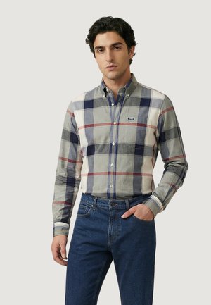 Man wearing a long-sleeve button-up shirt with blue, gray, and red plaid pattern and blue jeans, standing with one hand in pocket.
