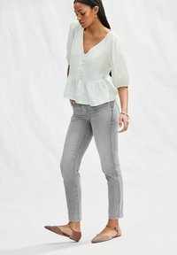 Blusa blanca texturizada con un escote en V profundo y mangas abullonadas; combinada con jeans ajustados grises y zapatos planos de color marrón claro.