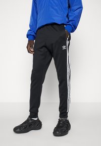 Černé sportovní kalhoty s bílými pruhy po stranách, s elastickým pasem a hladkou texturou, doplněné o černé tenisky s texturou.