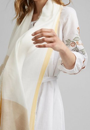 Femme portant une robe blanche avec des manches brodées de fleurs et tenant une grande écharpe blanche aux bords beige et jaune.