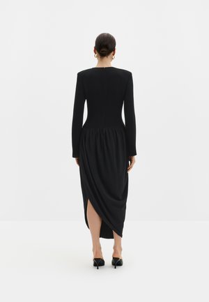 Femme avec les cheveux en chignon portant une longue robe noire à manches longues, jupe froncée, fente dans le dos et chaussures à talons noirs, debout de dos.