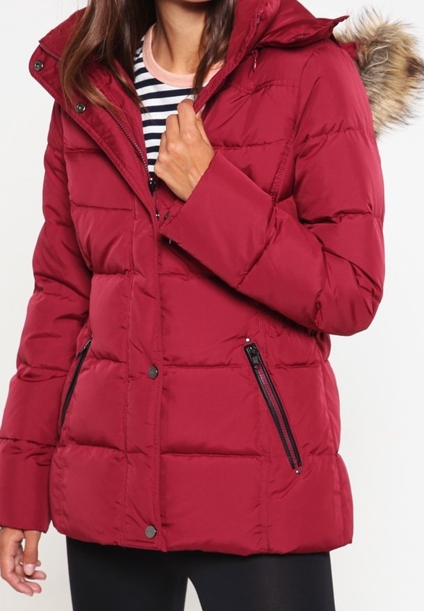 Giacca imbottita rossa con cappuccio con pelliccia, dotata di tasche con zip, bottoni a pressione e un design trapuntato. Indossata sopra una maglietta a righe.