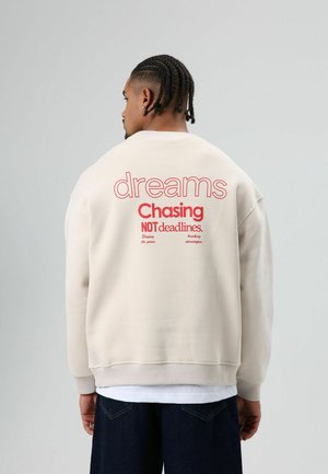 Beige sweatshirt met rode tekst op de achterkant: "dreams Chasing NOT deadlines." Oversized pasvorm met geribde boorden en zoom.